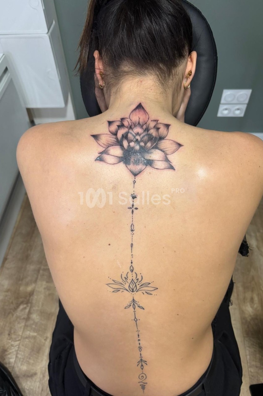 Femme de dos avec un tatouage détaillé représentant une fleur de lotus et des motifs géométriques le long de la colonne…