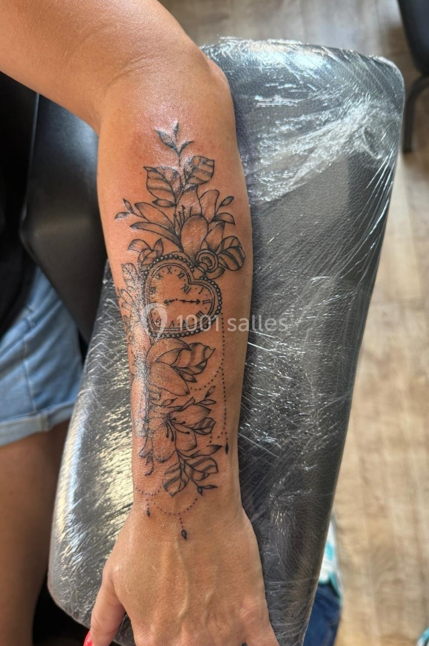 Tatouage sur l'avant-bras représentant un cœur en forme de montre entouré de fleurs et de feuilles.