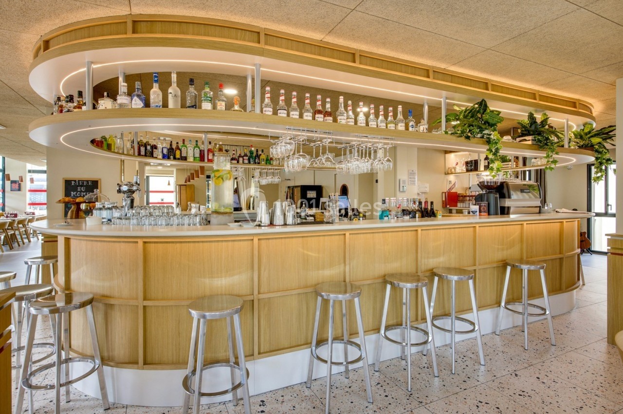 Bar moderne en bois clair avec étagères garnies de bouteilles, tabourets métalliques et plantes décoratives.