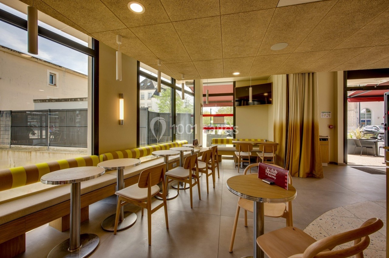 Salle lumineuse avec tables et chaises en bois, banquettes jaunes, grandes fenêtres et décoration moderne.