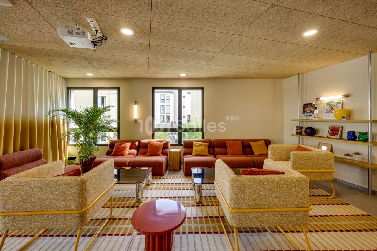 Espace salon lumineux avec fauteuils et canapés colorés, tapis à motifs, étagères décorées et plantes.