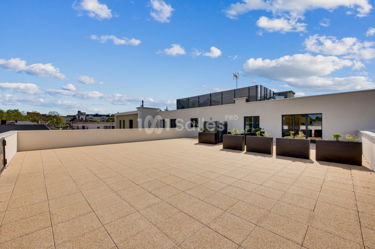 Terrasse spacieuse avec sol en dalles beige, jardinières modernes et vue dégagée sous un ciel partiellement nuageux.
