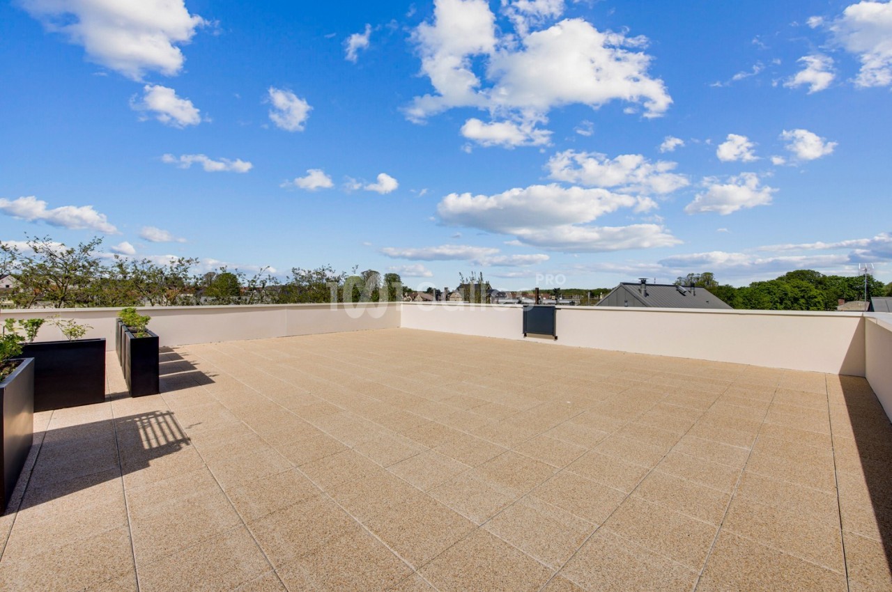 Terrasse spacieuse avec sol en dalles beige, bordée de jardinières, sous un ciel bleu parsemé de nuages.