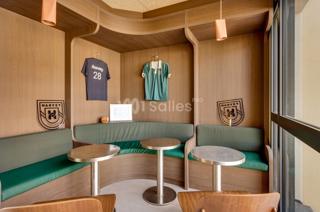 Coin salon avec banquettes vertes, tables rondes, maillots de football accrochés au mur et lumière naturelle.