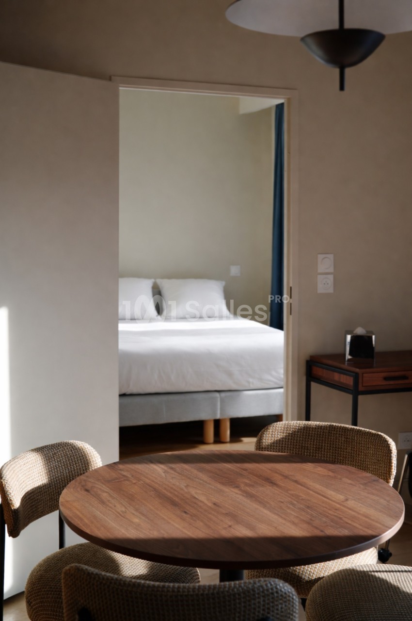 Vue d'une chambre avec un lit double à travers une porte ouverte, table ronde en bois et chaises au premier plan.