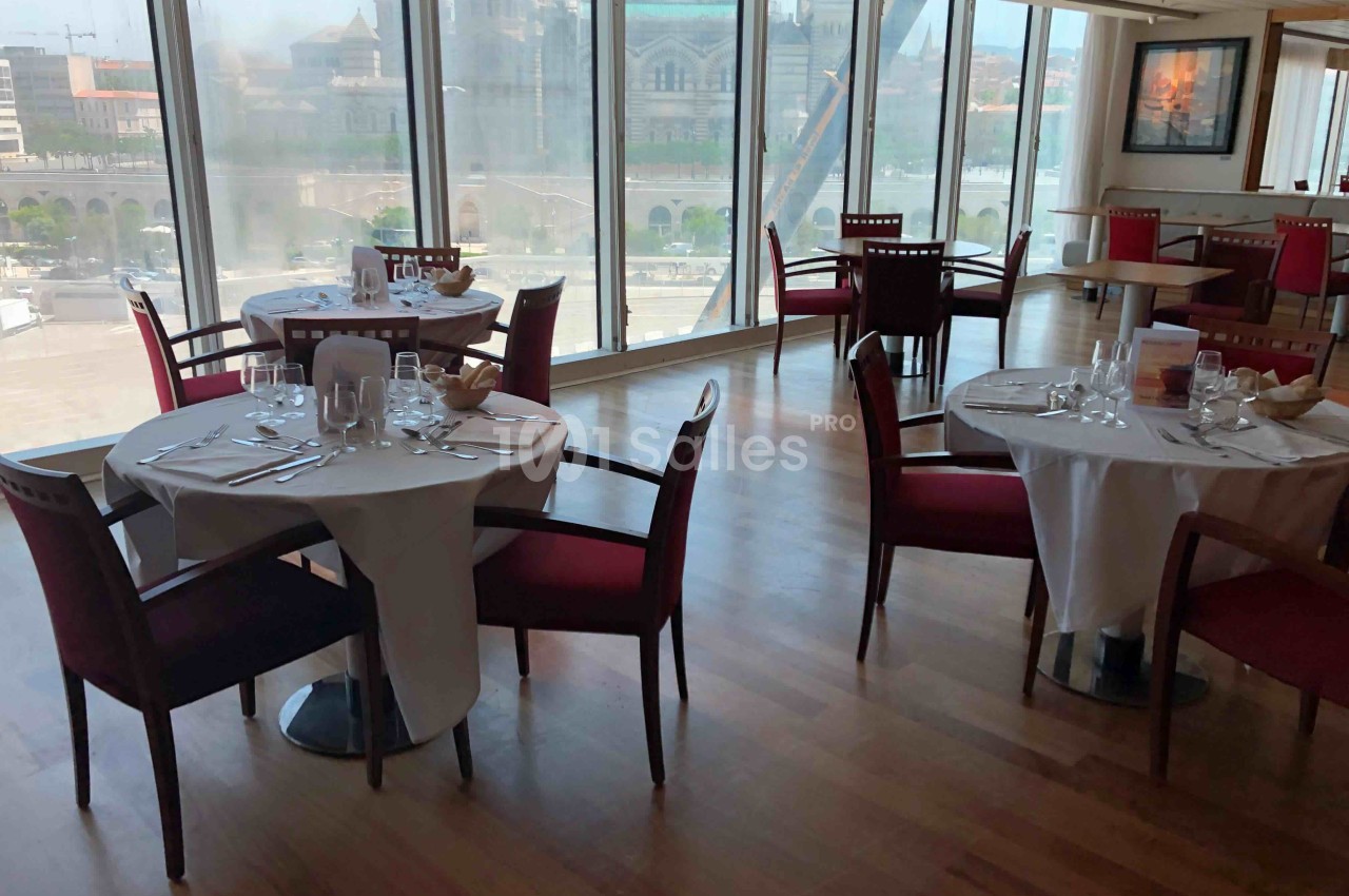 Salle de restaurant lumineuse avec tables dressées, chaises rouges et vue sur un paysage urbain à travers de grandes baies…