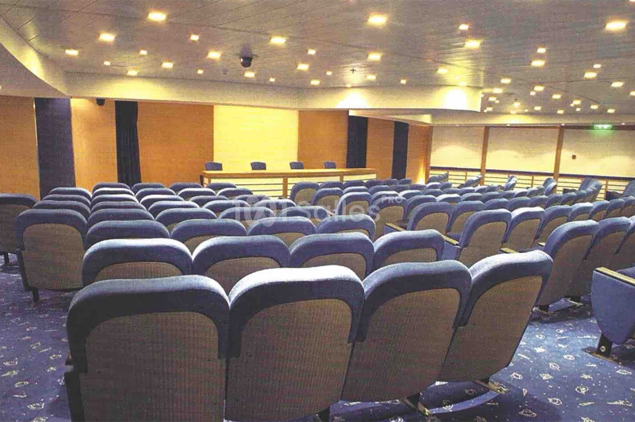Salle de conférence vide avec rangées de sièges bleus et une estrade équipée de micros en arrière-plan.