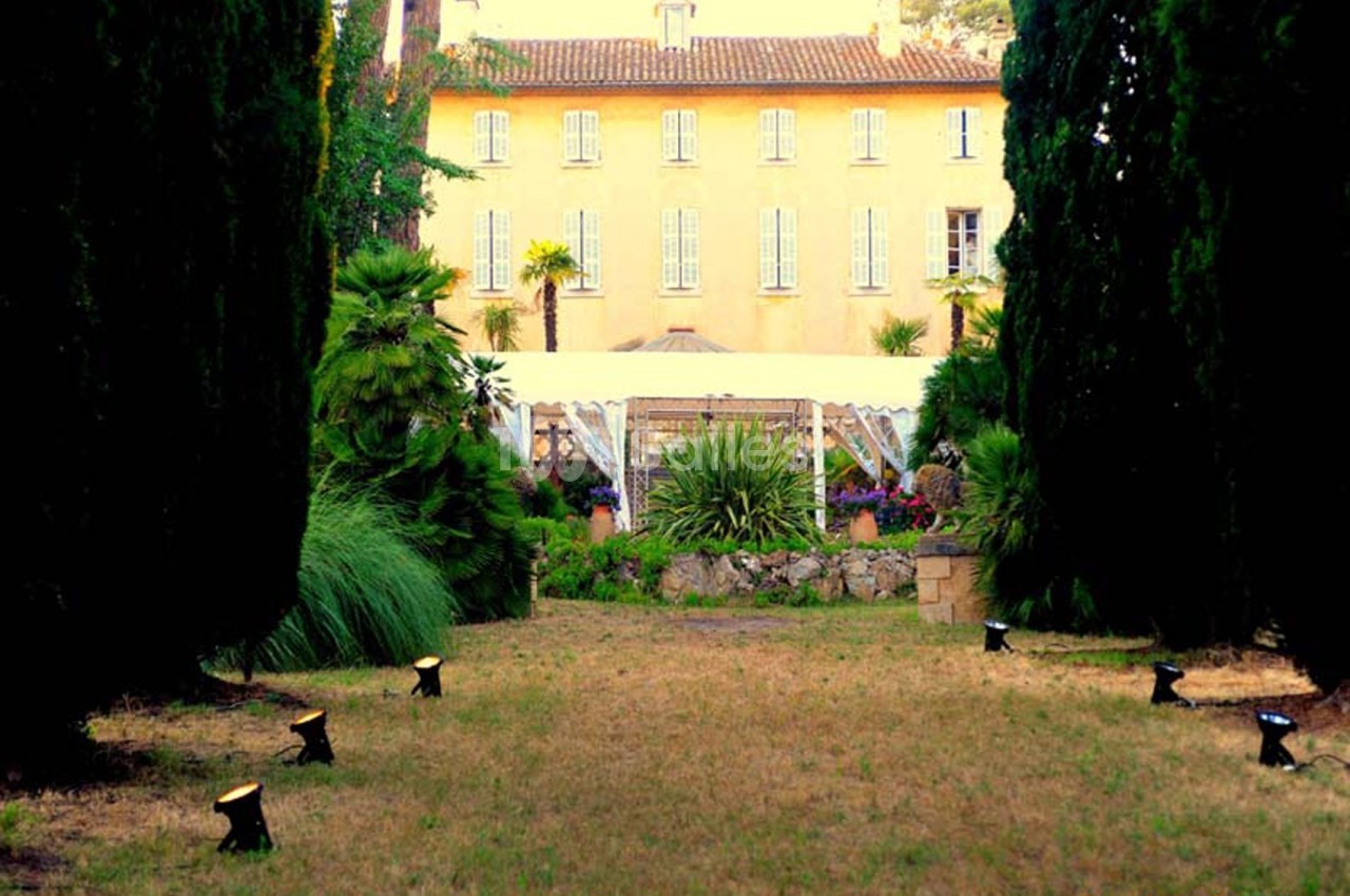 Allée bordée d'arbres menant à une grande maison jaune entourée de végétation et d'un jardin aménagé.