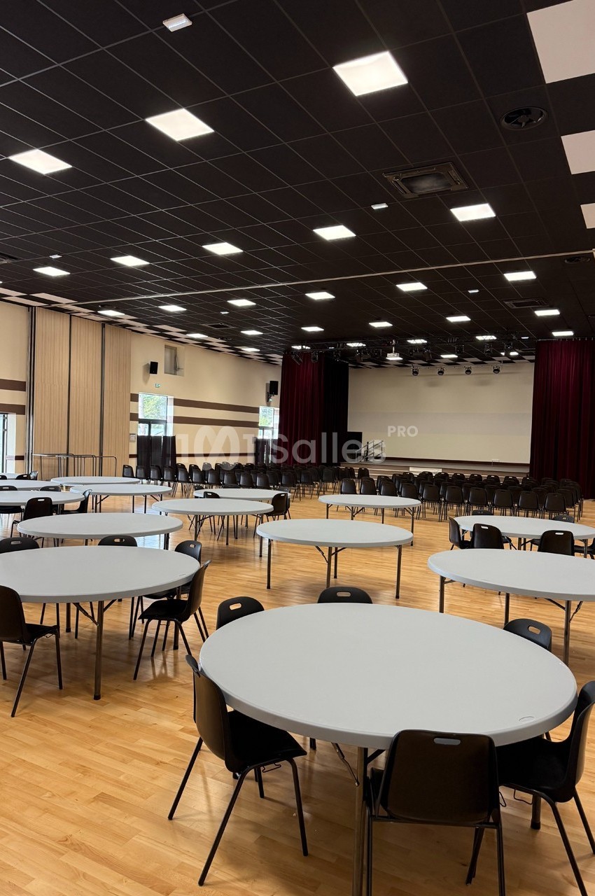 Salle polyvalente avec parquet, tables rondes, chaises noires, scène avec rideaux rouges et plafond éclairé.