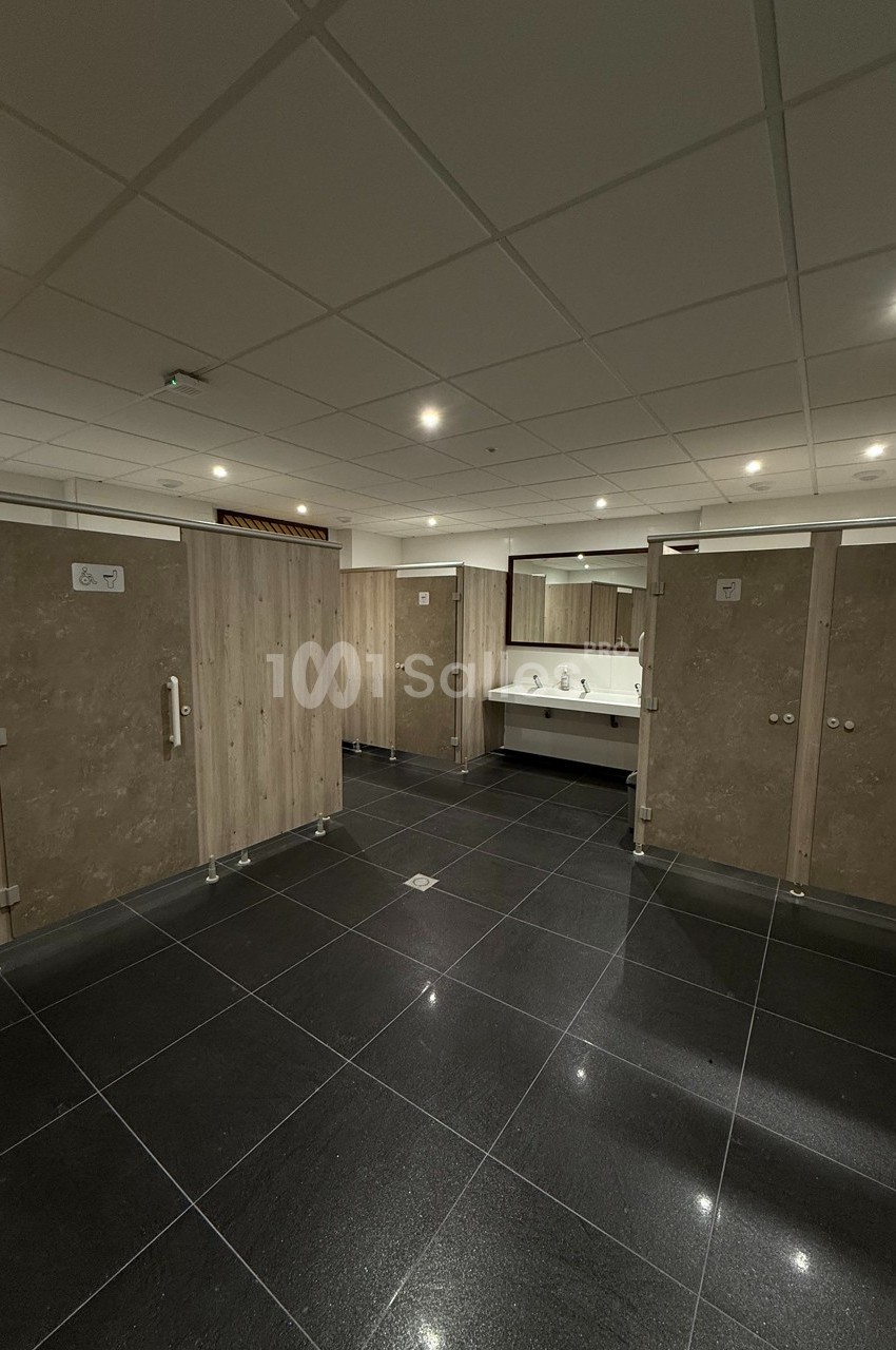 Vue d'un espace de toilettes publiques avec cabines fermées, lavabos et miroir, sol en carrelage noir.