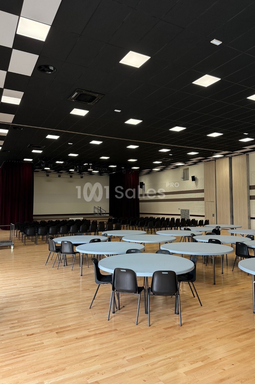 Salle polyvalente avec parquet, tables rondes, chaises noires, scène avec rideaux rouges et plafond à éclairage intégré.