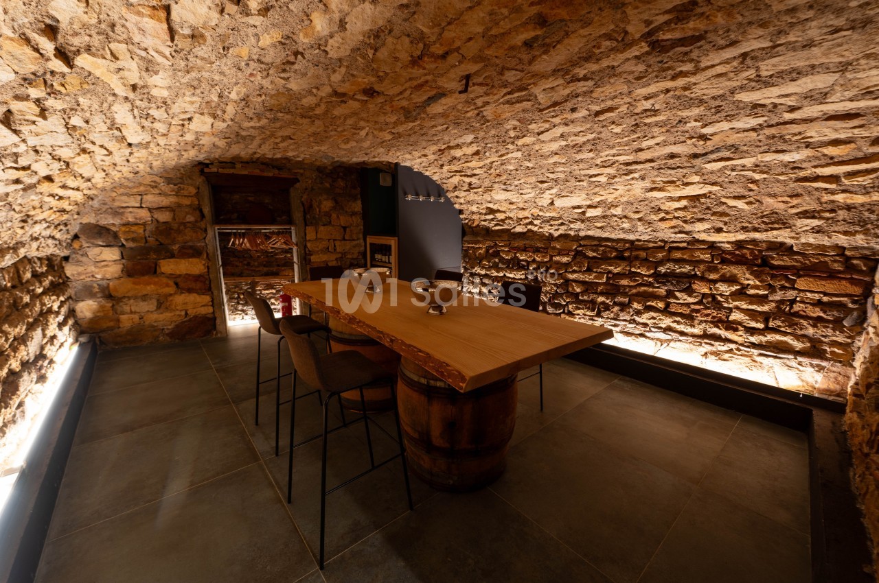 Salle voûtée en pierre avec une table en bois sur un tonneau, entourée de chaises, éclairage tamisé.