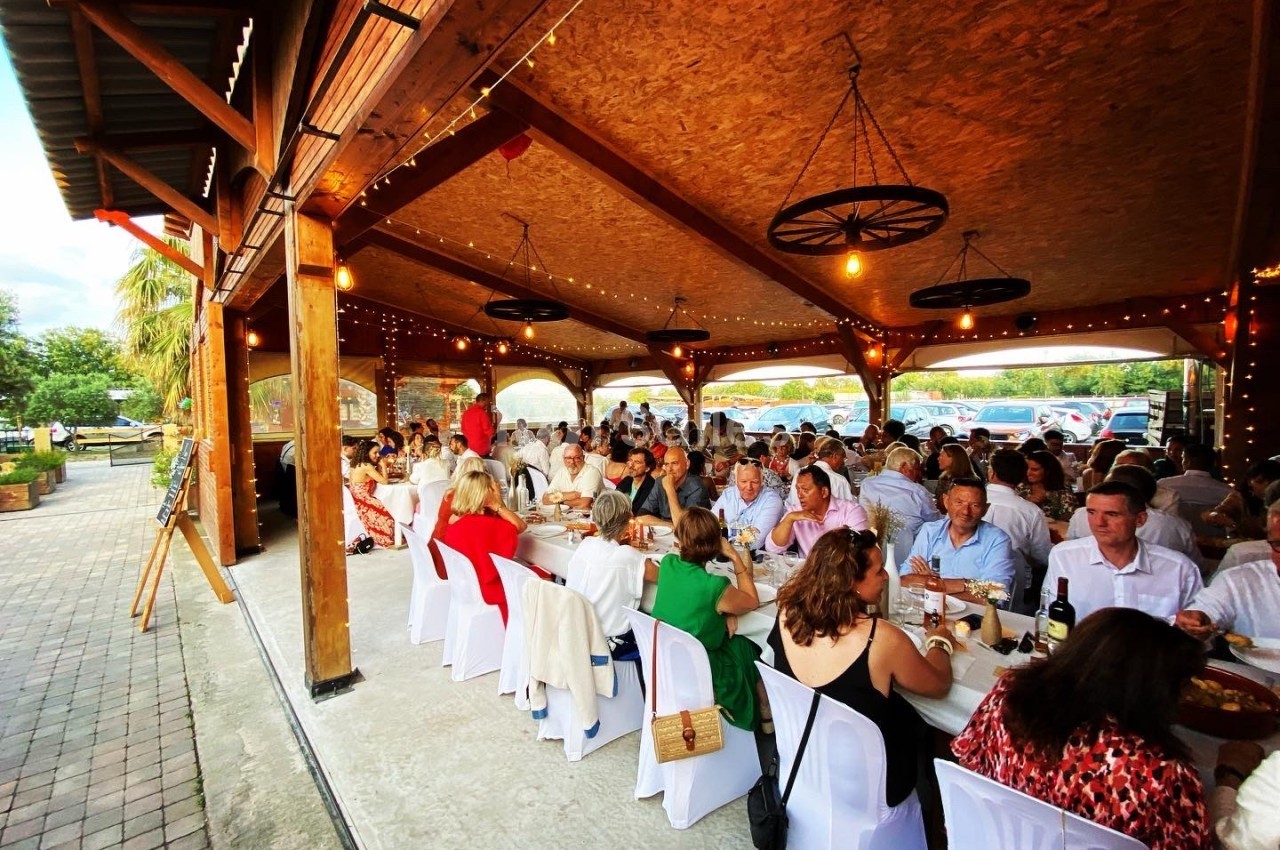 Des convives assis à de longues tables sous un auvent en bois, décoré de lumières suspendues, lors d'un événement festif.