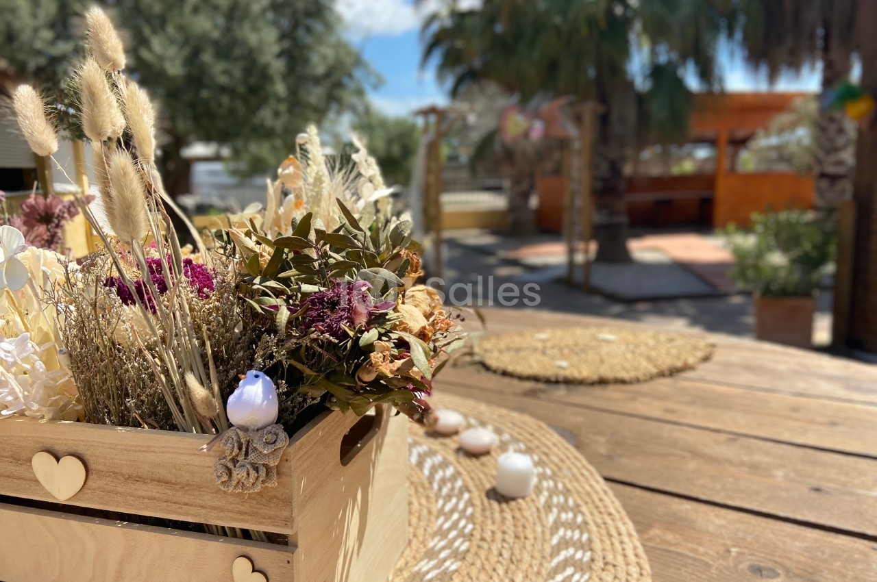 Centre de table en bois avec des fleurs séchées, posé sur une table en bois dans un espace extérieur ensoleillé.