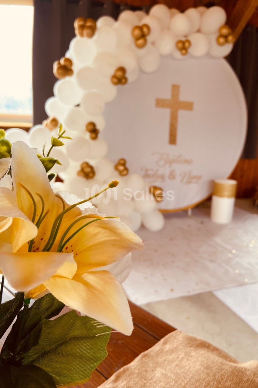 Arrangement floral au premier plan avec en arrière-plan une décoration de baptême composée de ballons blancs et dorés.