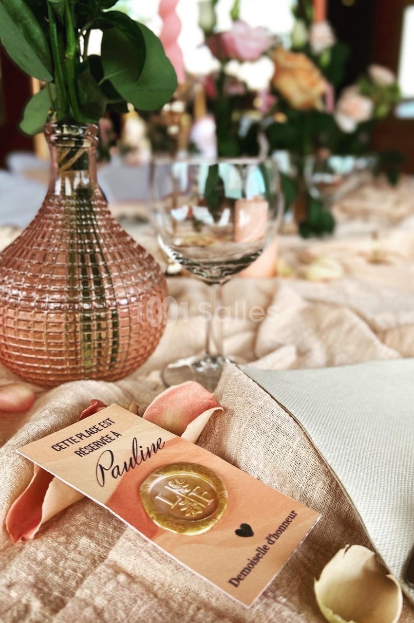Table décorée avec des fleurs, verrerie et un marque-place personnalisé avec un sceau doré et le prénom ’Pauline’.