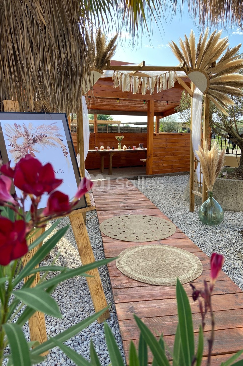 Allée en bois décorée de tapis ronds menant à une pergola ornée de pampas, avec fleurs rouges au premier plan.