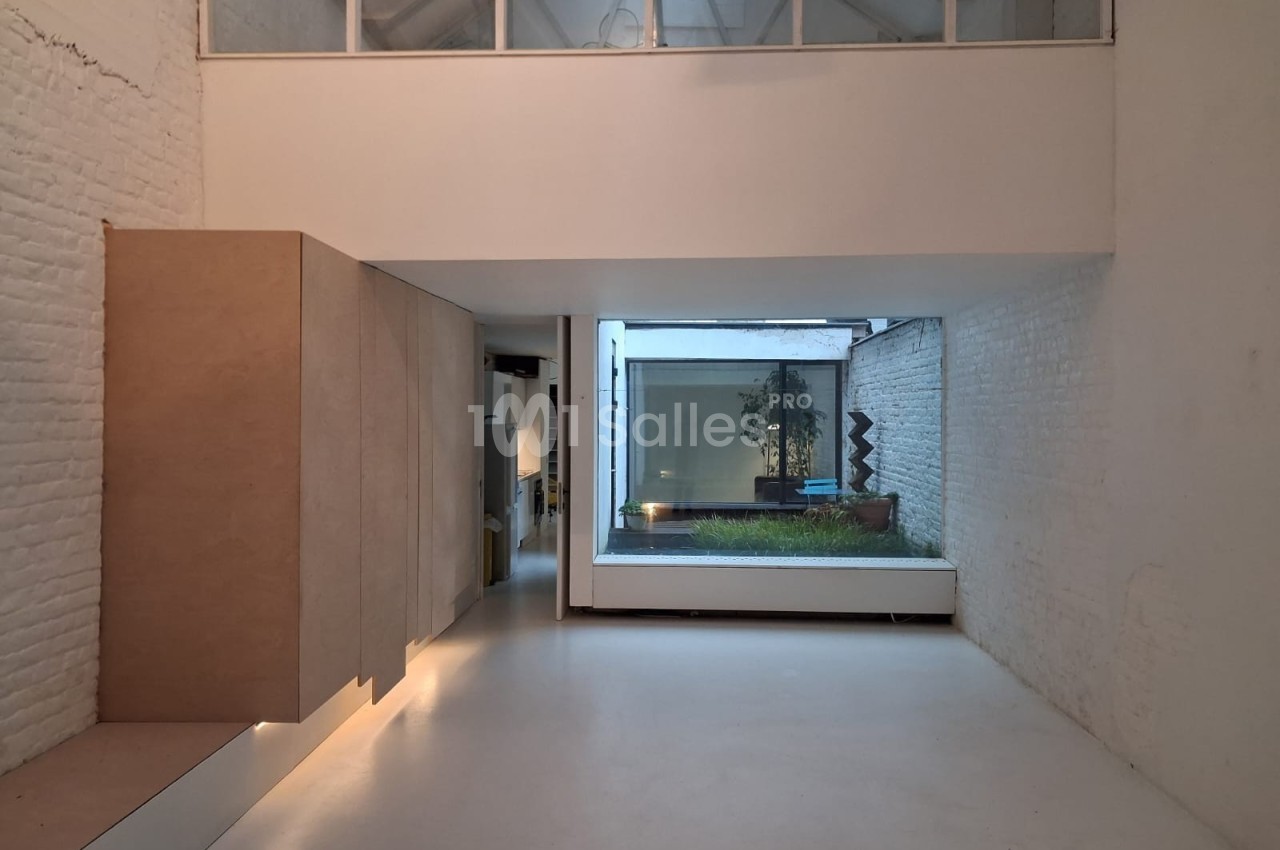 Intérieur minimaliste avec murs blancs, grandes fenêtres, mobilier épuré et vue sur une cour avec végétation.