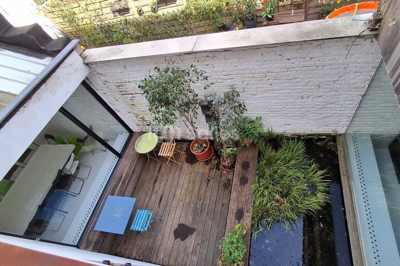 Vue d'une petite terrasse en bois avec des plantes en pots, une table, des chaises et un bassin bordé de végétation.