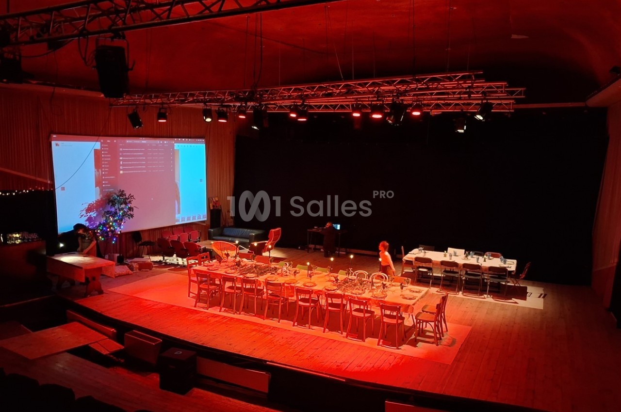 Salle de spectacle éclairée en rouge avec une grande table dressée, des chaises, un écran projeté et des équipements…