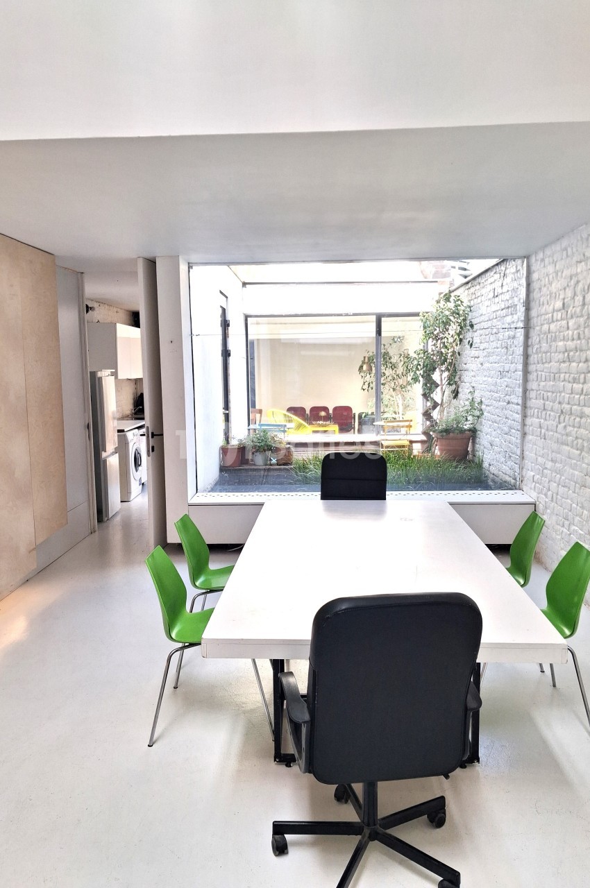 Salle de réunion lumineuse avec table blanche, chaises vertes et vue sur une cour intérieure avec plantes.
