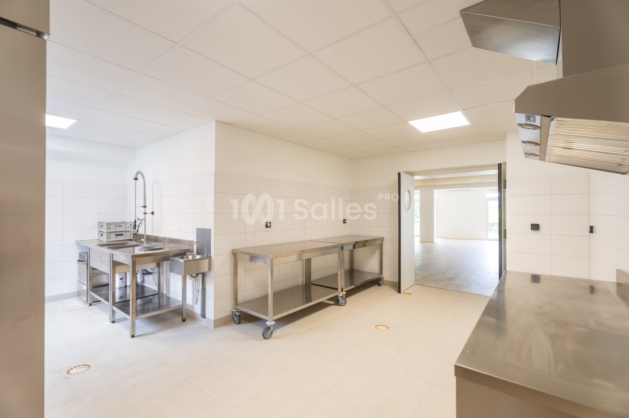 Cuisine professionnelle vide avec plans de travail en inox, évier, carrelage blanc et éclairage au plafond.