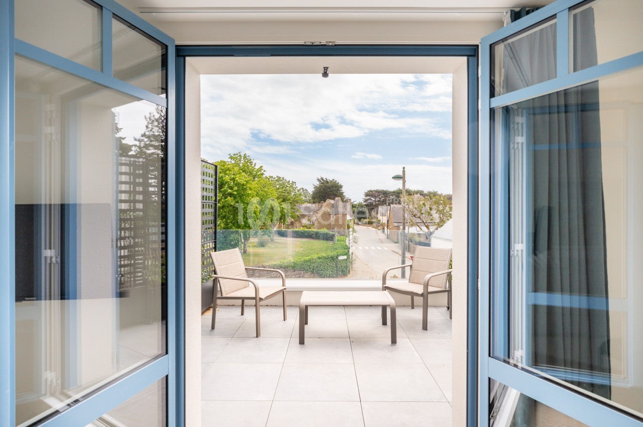 Vue depuis une pièce sur une terrasse aménagée avec des fauteuils et une table, donnant sur un jardin et une rue.