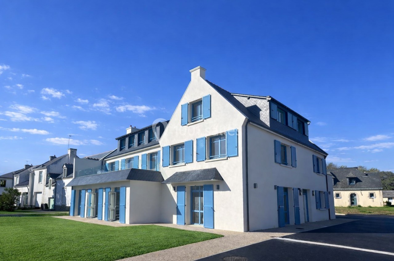 Maison blanche à trois étages avec volets bleus, située dans un quartier résidentiel sous un ciel dégagé.