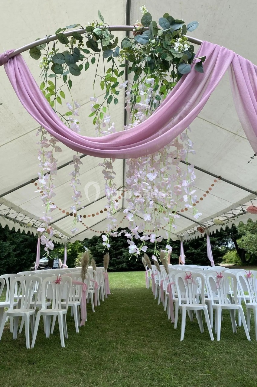 Tente blanche décorée de guirlandes florales roses et vertes, avec des rangées de chaises blanches sur une pelouse.