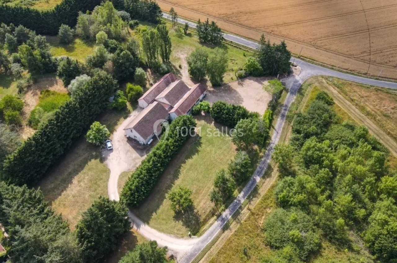 Vue aérienne d'une maison entourée de jardins, d'arbres et de champs, bordée par une route sinueuse.