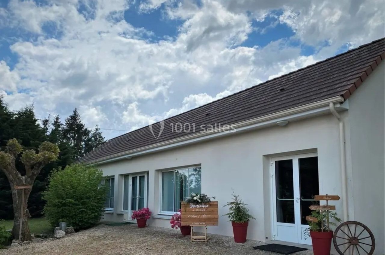 Façade d'une maison blanche avec des grandes fenêtres, des pots de fleurs et un panneau en bois devant.