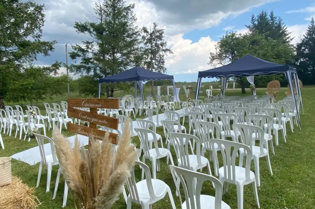 Chaises blanches disposées en rangées sous des tonnelles bleues dans un espace extérieur verdoyant, avec un panneau en bois.