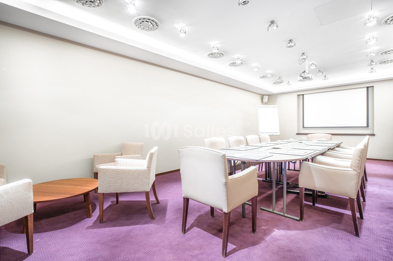 Salle de réunion lumineuse avec table rectangulaire, chaises blanches, moquette violette et écran de projection.