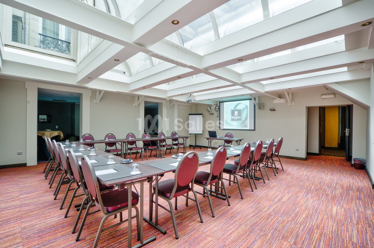 Salle de réunion lumineuse avec tables disposées en U, chaises rouges, projecteur et tableau blanc.