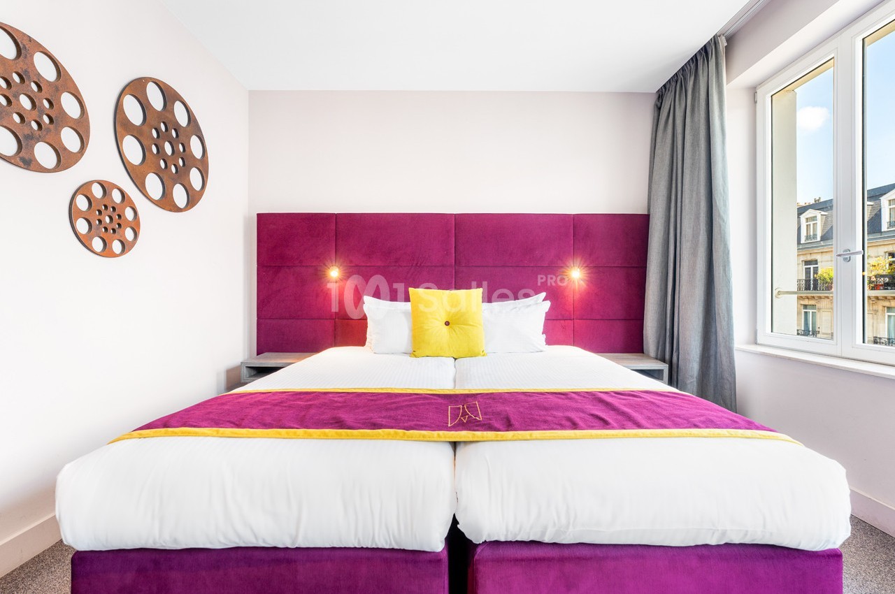 Lit double avec tête de lit violette, coussins colorés, décor mural circulaire et fenêtre donnant sur des bâtiments.