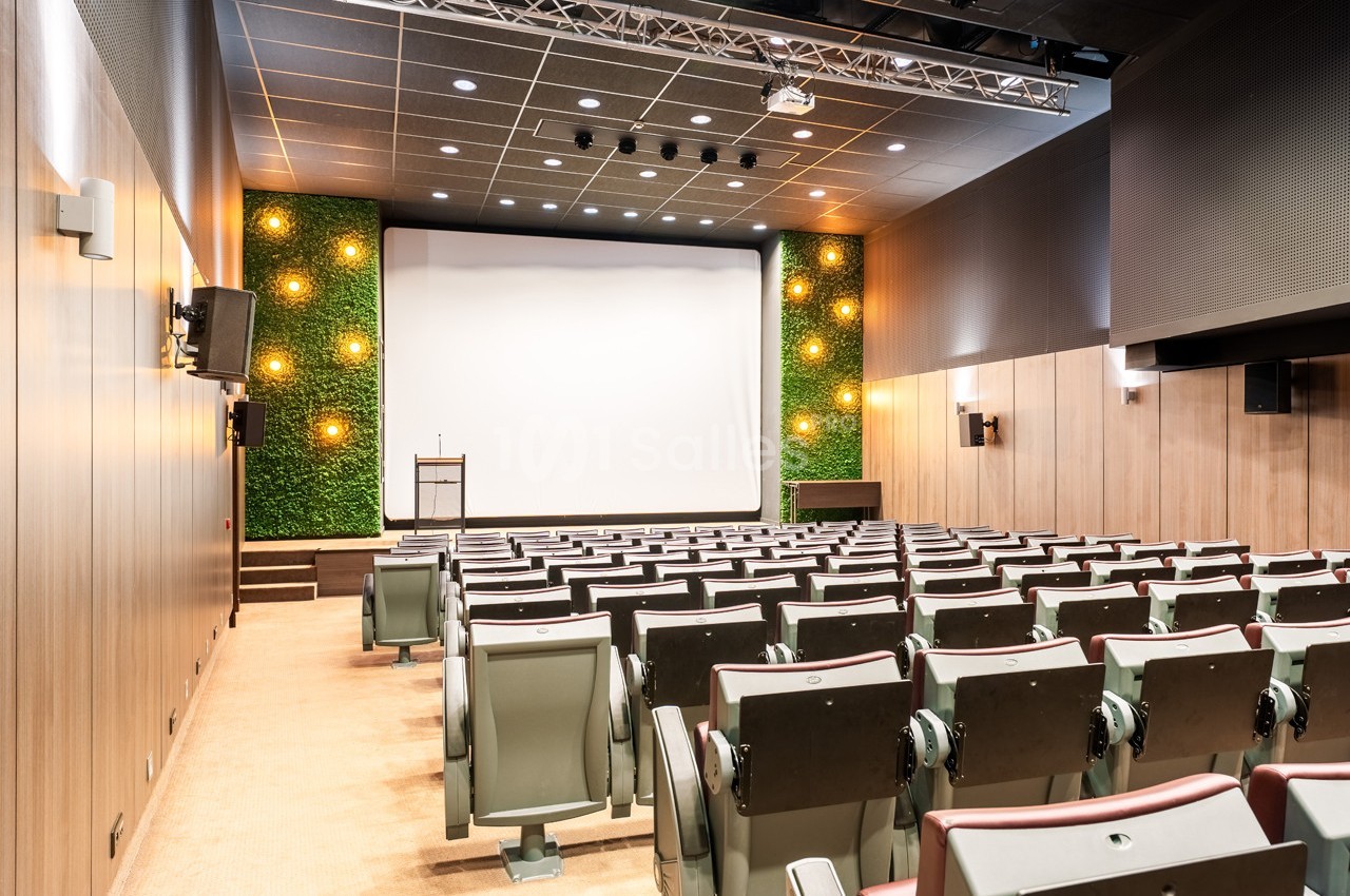 Salle de conférence moderne avec écran blanc, sièges alignés, murs en bois et décor végétal éclairé.