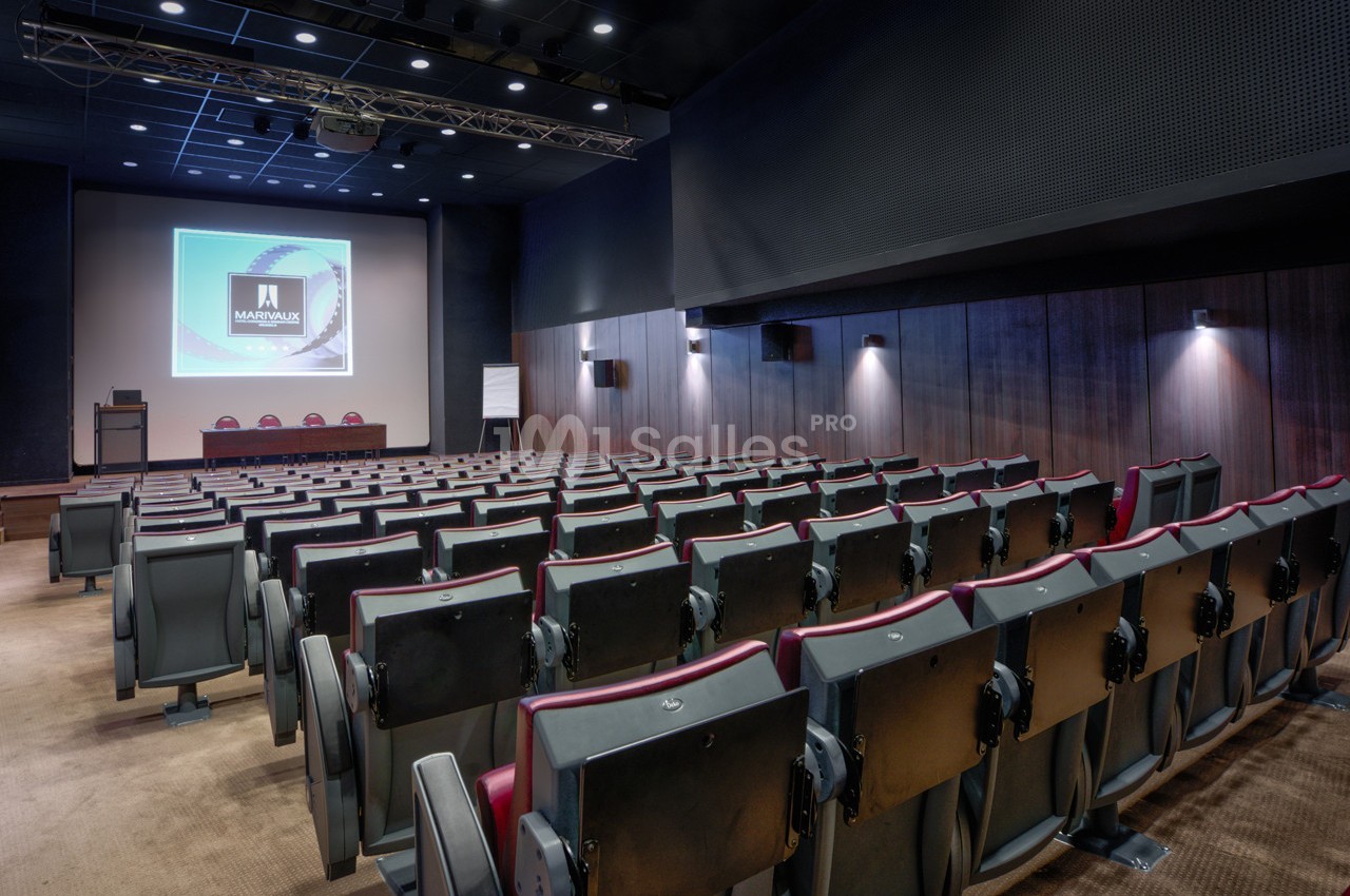 Salle de conférence vide avec rangées de sièges, écran de projection allumé et éclairage tamisé.