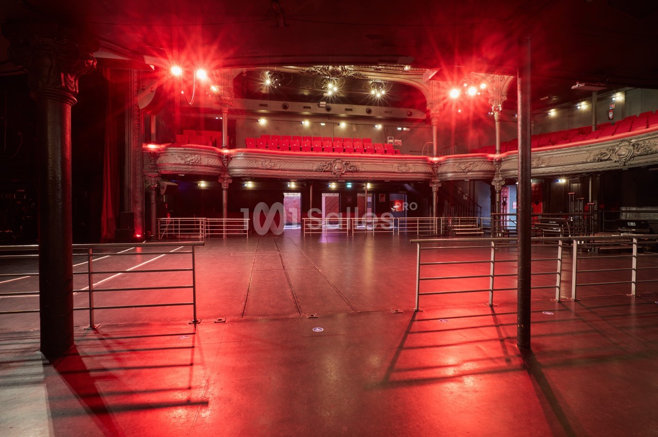 Salle de spectacle vide avec balustrades, scène et balcons ornés, éclairée par des lumières rouges.