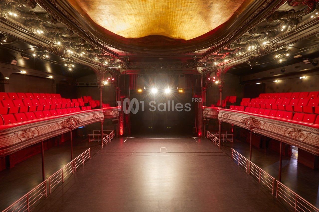 Salle de théâtre vide avec balcons ornés, sièges rouges et scène éclairée par des projecteurs.