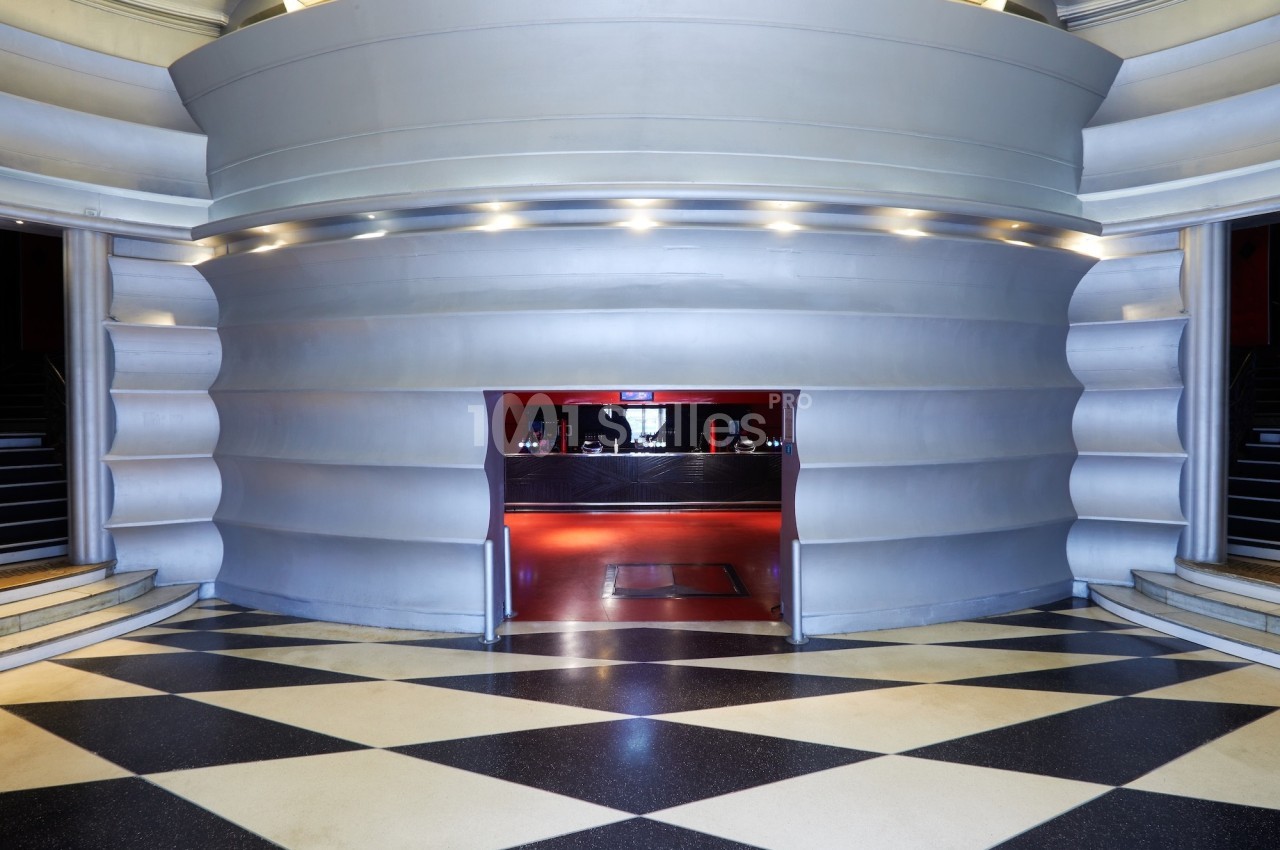 Entrée d'un hall moderne avec des murs métalliques incurvés, sol en damier et lumière rouge en arrière-plan.