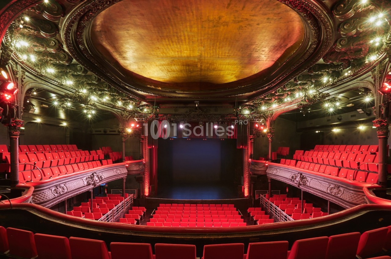 Salle de théâtre vide avec sièges rouges, balcon orné et plafond décoré, éclairée par des lumières tamisées.