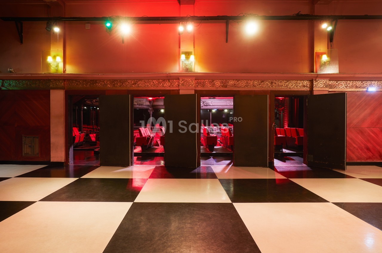 Salle de cinéma avec sol en damier, portes ouvertes sur des rangées de sièges rouges sous un éclairage tamisé.