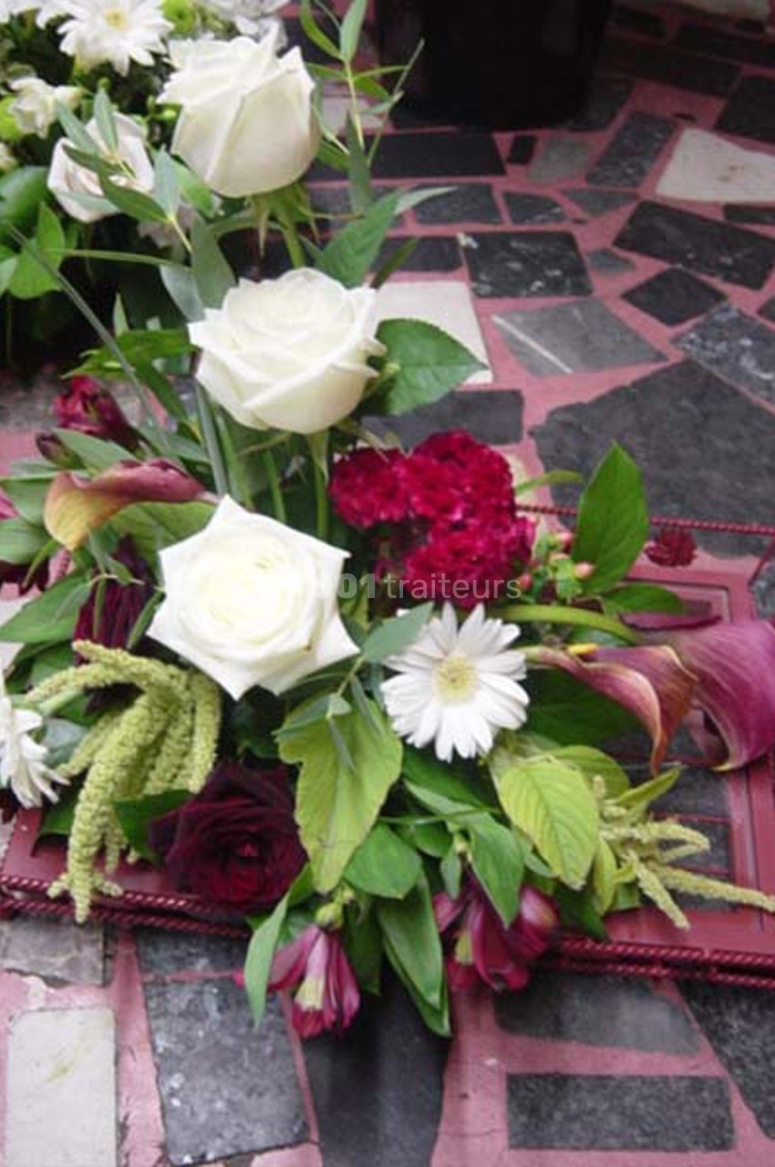 Arrangement floral avec roses blanches, fleurs rouges et feuillage vert, posé sur un cadre rouge sur un sol carrelé.