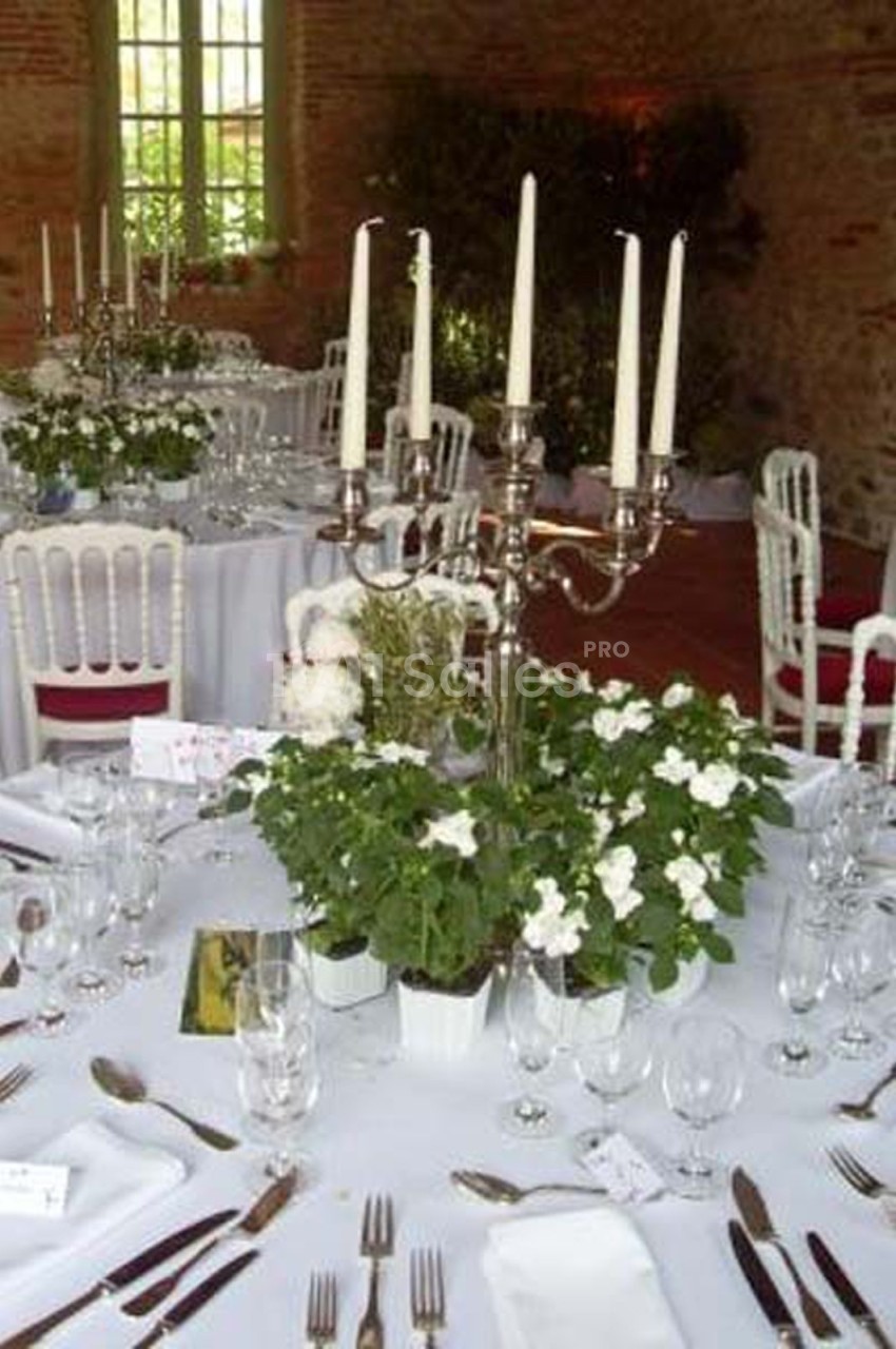 Tables élégamment dressées avec nappes blanches, chandeliers et arrangements floraux dans une salle lumineuse en pierre.