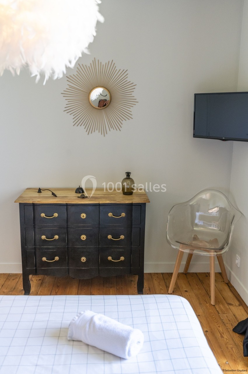 Chambre avec commode noire, chaise transparente, miroir mural décoratif et télévision fixée au mur.
