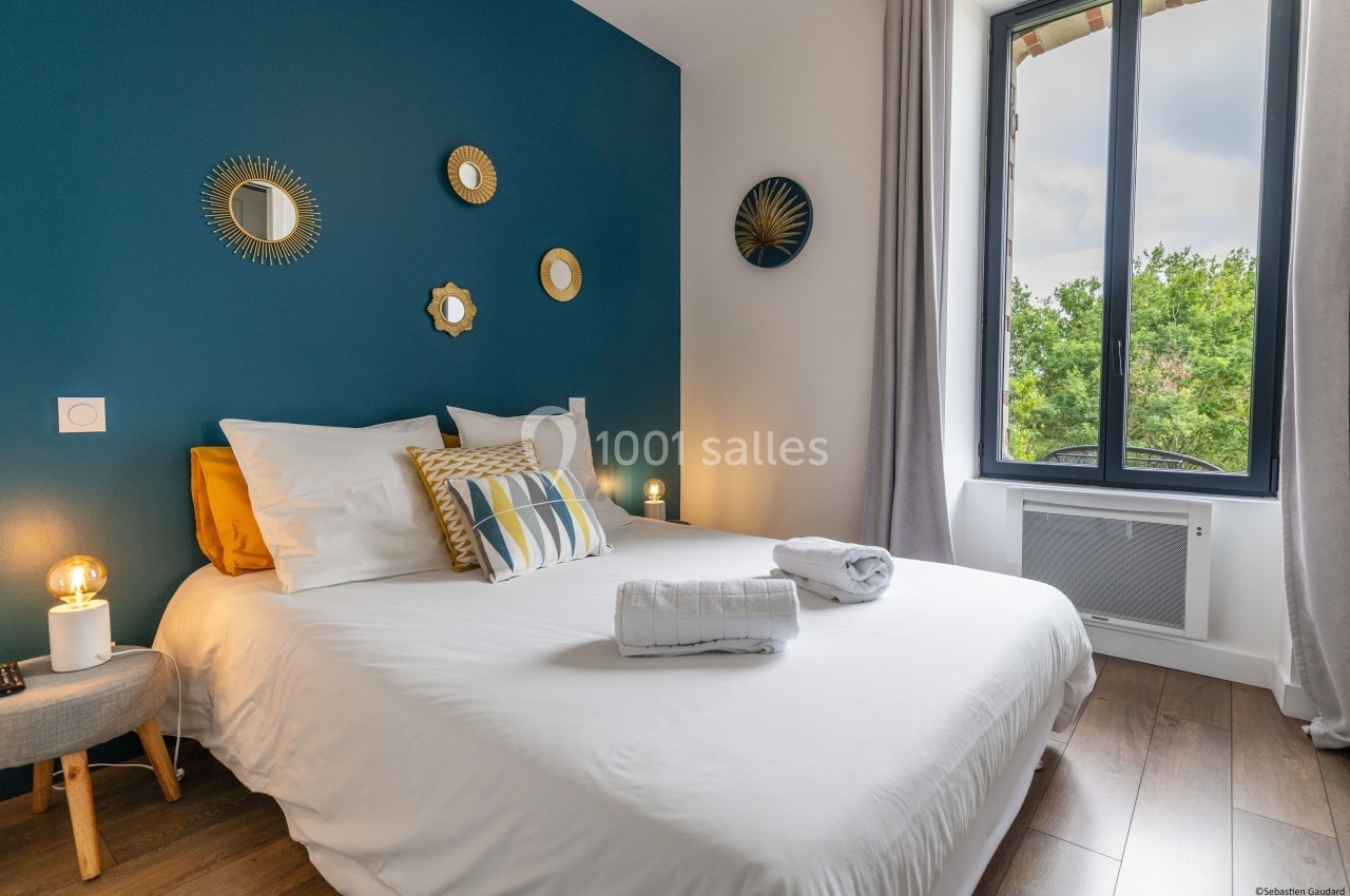 Chambre lumineuse avec lit double, mur bleu décoré de miroirs, fenêtre donnant sur des arbres et sol en parquet.