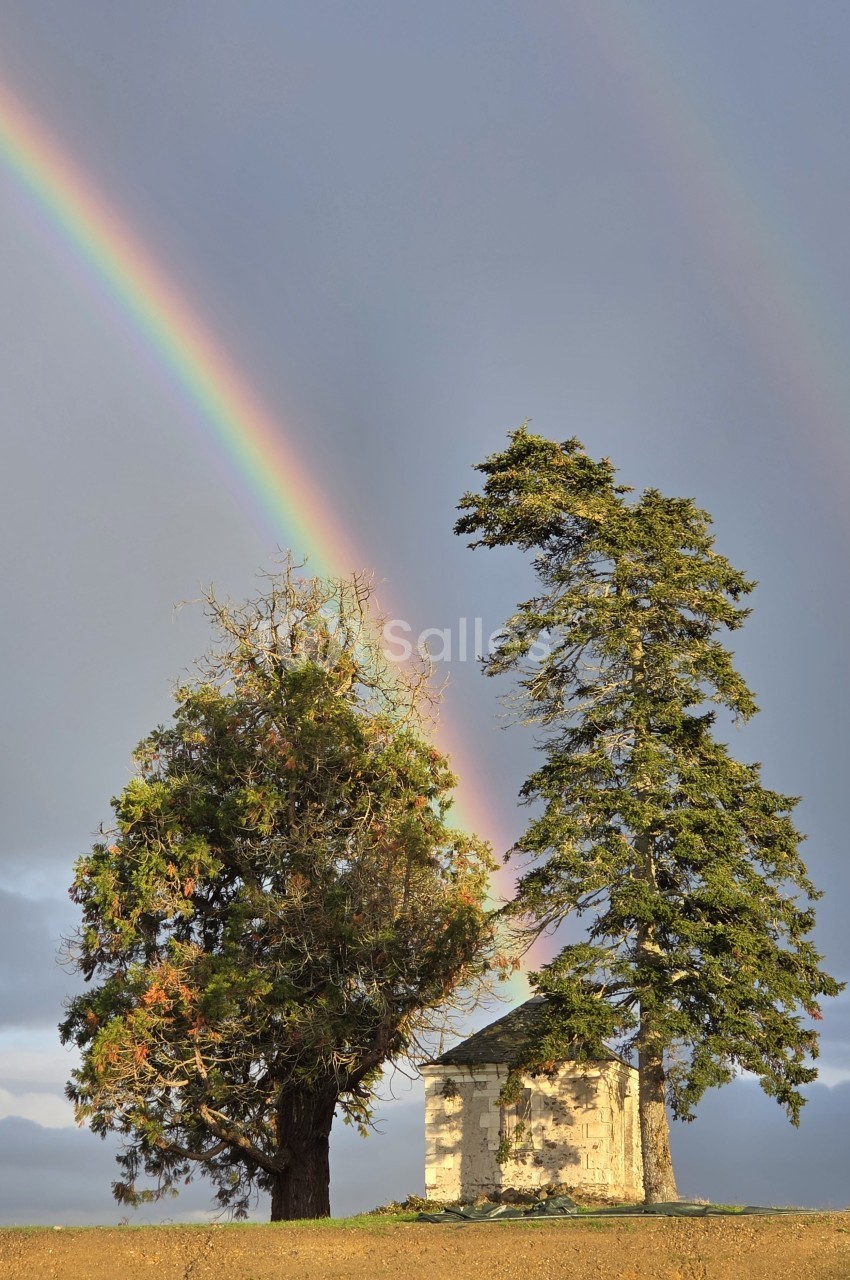 Un petit bâtiment en pierre entouré de deux arbres, avec un arc-en-ciel visible dans un ciel nuageux.