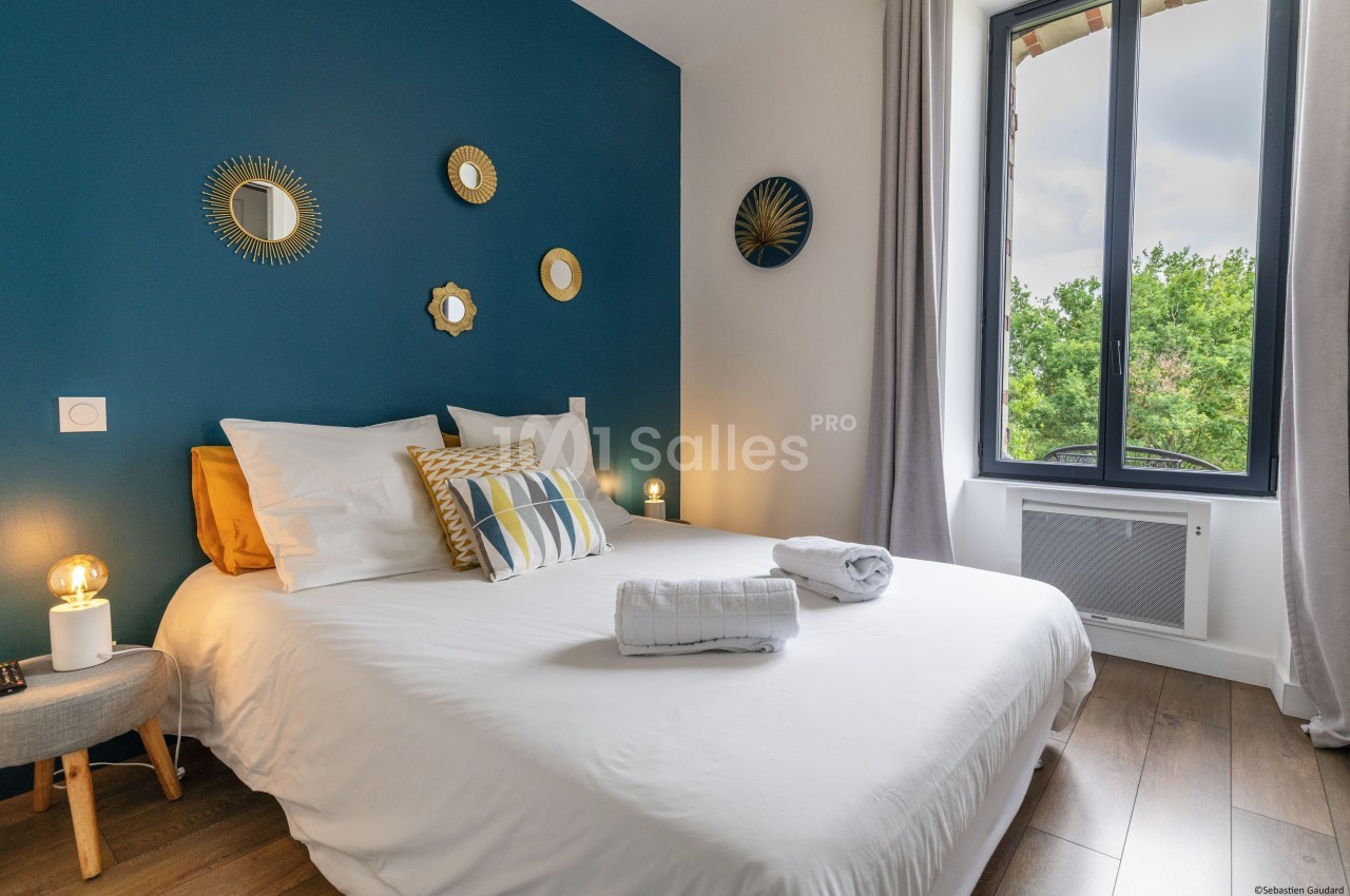 Chambre lumineuse avec lit double, mur bleu décoré de miroirs, fenêtre donnant sur des arbres et parquet au sol.
