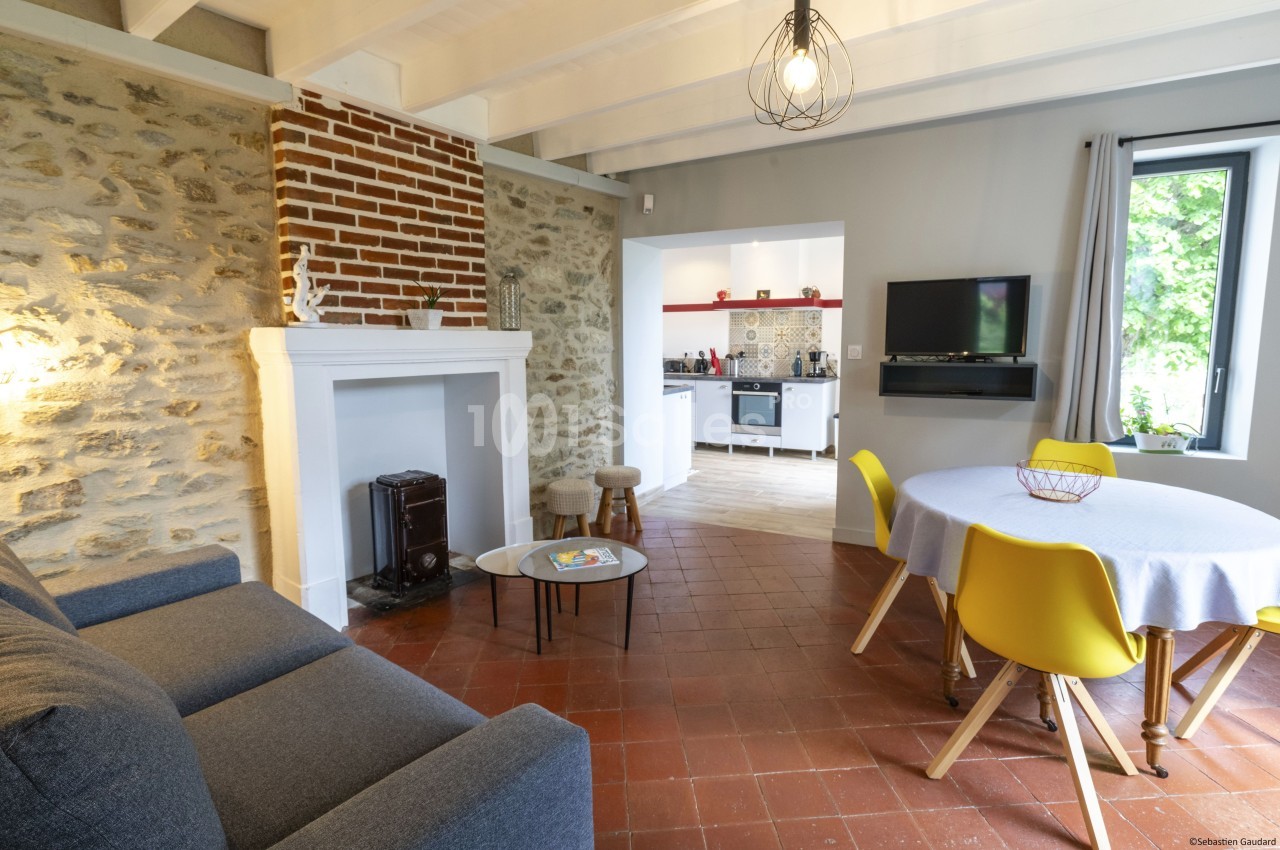 Salon lumineux avec murs en pierre, cheminée, canapé, table ronde avec chaises jaunes et vue sur une cuisine ouverte.