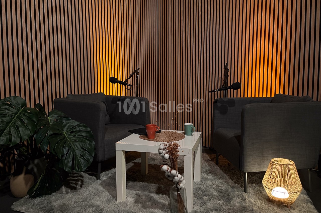 Deux fauteuils gris autour d'une table blanche avec des micros, dans un studio cosy éclairé par des lumières chaudes.